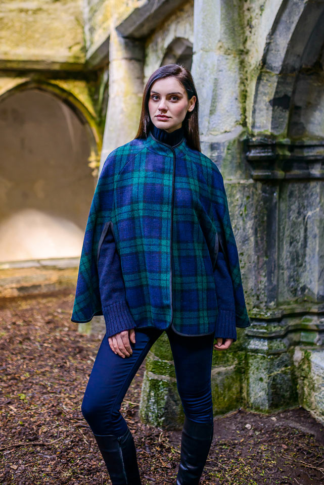 A woman in blue and green plaid cape standing next to an old stone wall.