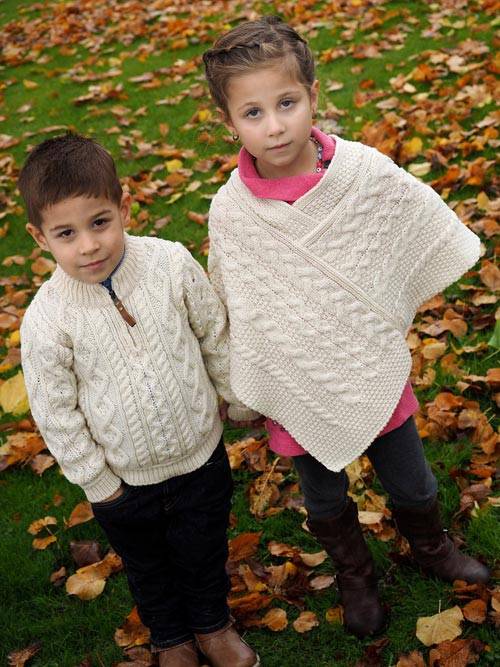 Two children in sweaters standing on a leaf covered lawn.