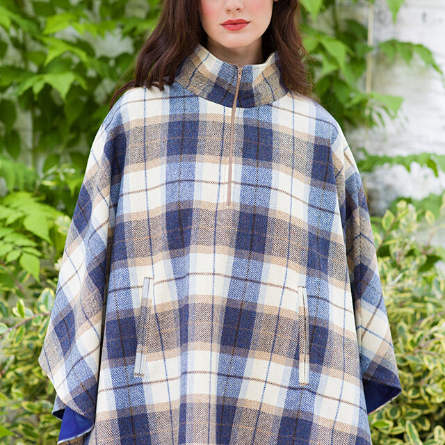 A woman in plaid poncho standing next to bushes.